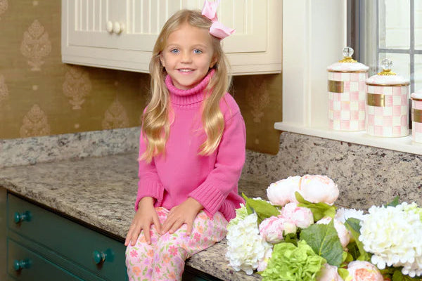 Young girl in pink sweater and polka dot pants sitting on a kitchen counter with flowers.
