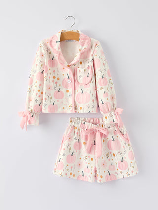 A two-piece children's pajama set hanging on a wooden hanger against a light gray background. The set includes a long-sleeve button-front top and matching shorts, both in a cream base fabric with a pink pumpkin and floral print, featuring pink ruffle trim, a button placket, and side bows.