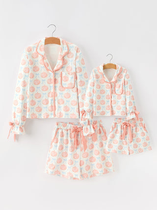 A matching pajama set for adults and children, featuring long-sleeve button-front shirts and shorts with a white background and a repeating peach pumpkin and bow pattern, displayed on wooden hangers against a light gray background.