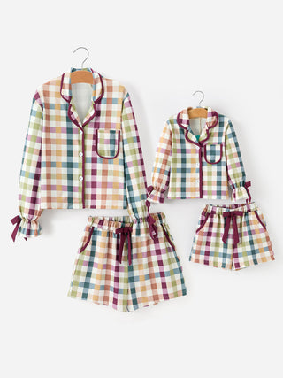 A matching pajama set for two sizes, featuring long-sleeve button-front shirts and shorts with a multicolor plaid pattern and dark plum trim, displayed on hangers against a plain white background.