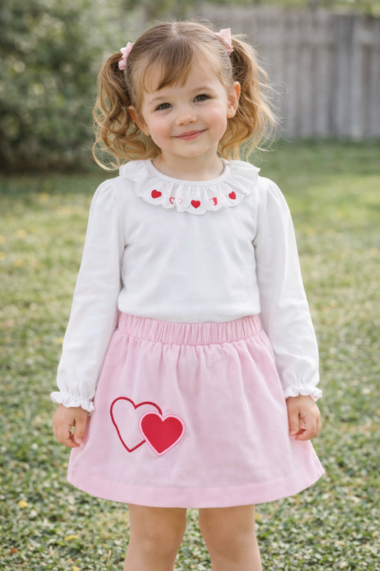Young girl wearing a white shirt with red heart designs and a pink skirt with red hearts outdoors.