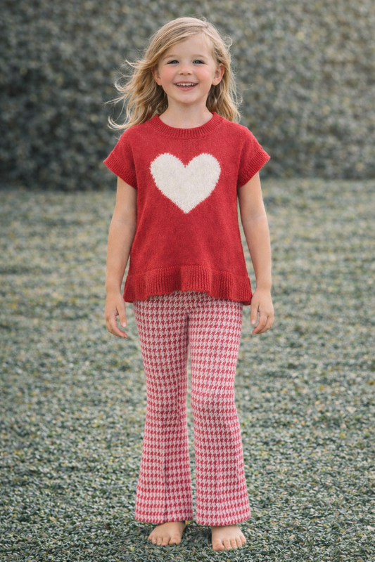 Young girl wearing a red sweater with a heart design and pink checkered pants standing on a textured surface.