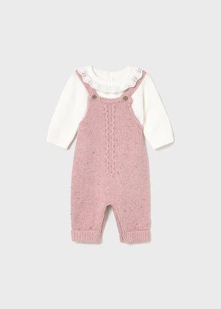 Pink knitted dungarees with a white shirt underneath on a light gray background